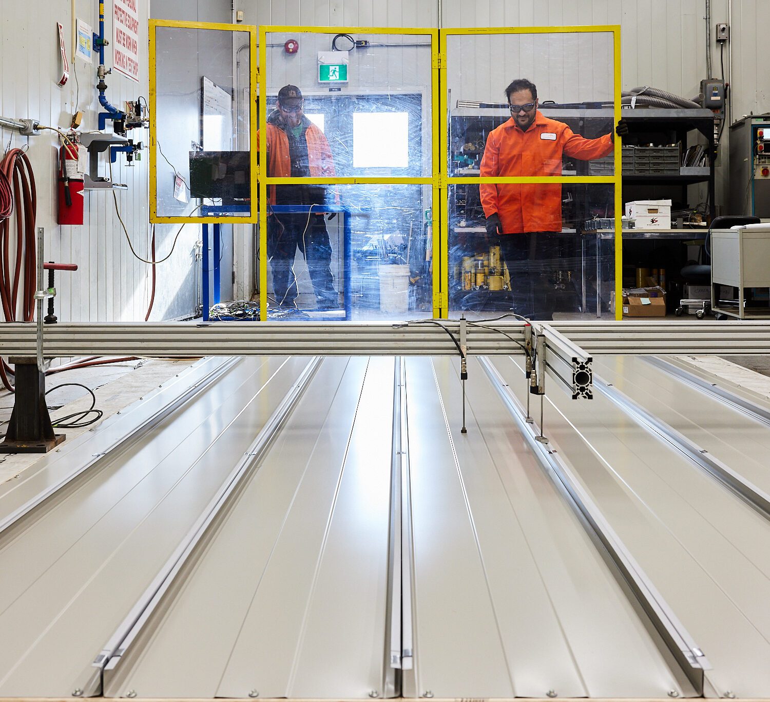 Two test lab staff wearing safety equipment, and standing behind protective glass in front of lab equipment.