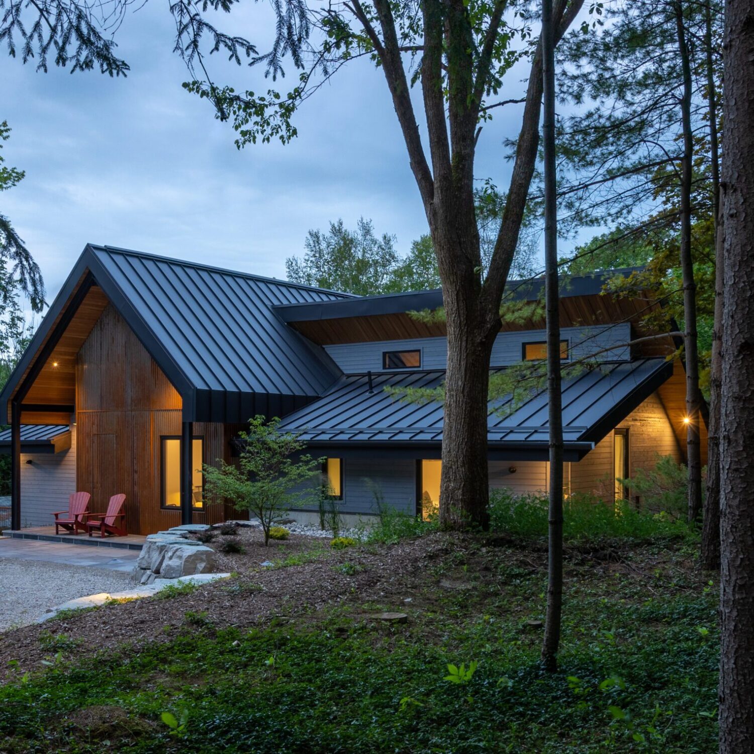 At dusk, the cabin is shown with rusted Indaten weathered steel exterior, viewed through forest trees