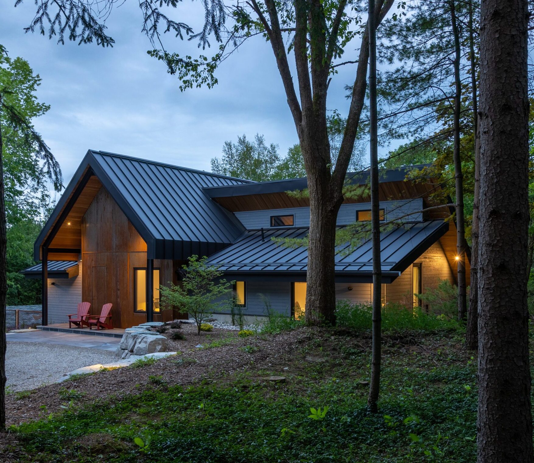 At dusk, the cabin is shown with rusted Indaten weathered steel exterior, viewed through forest trees